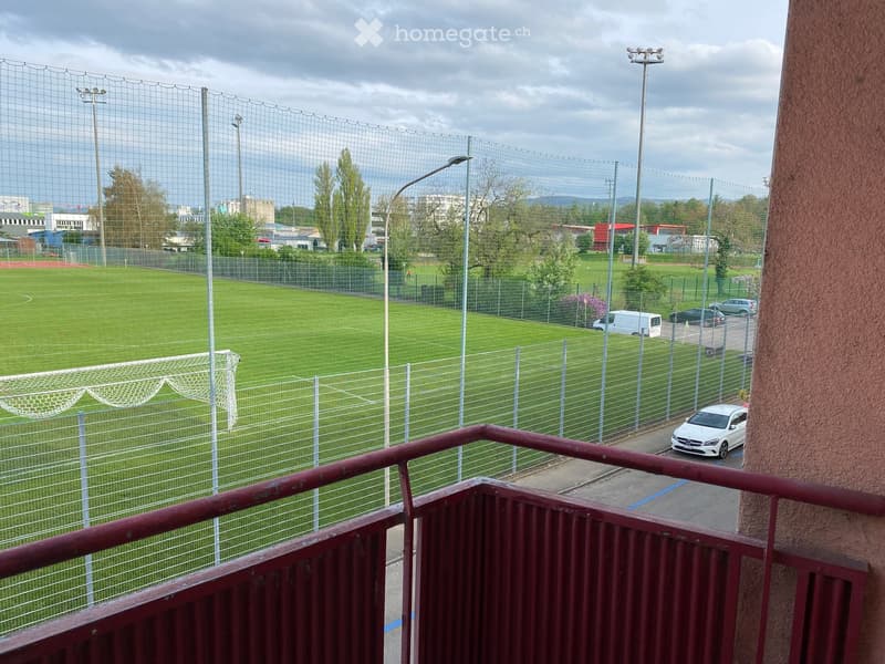 Balcony overlooking a fenced soccer field with goalposts, surrounded by trees and parked cars.