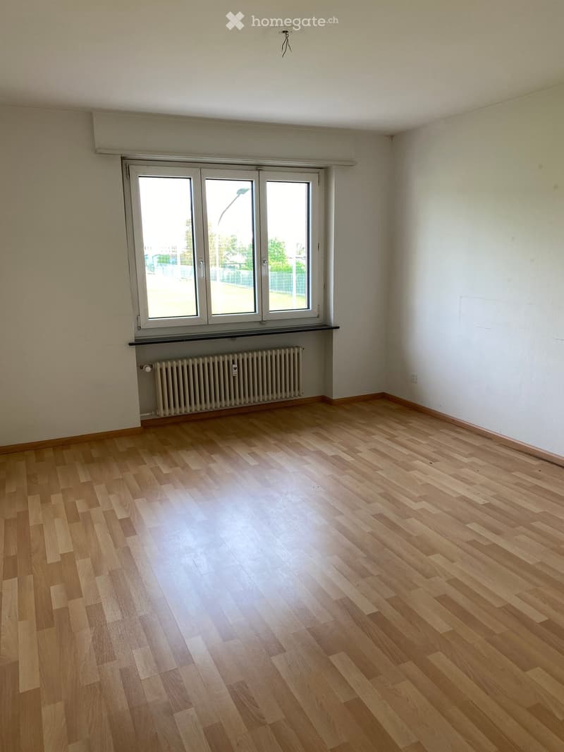 Empty room with wooden flooring, white walls, and a large window with a radiator below it.