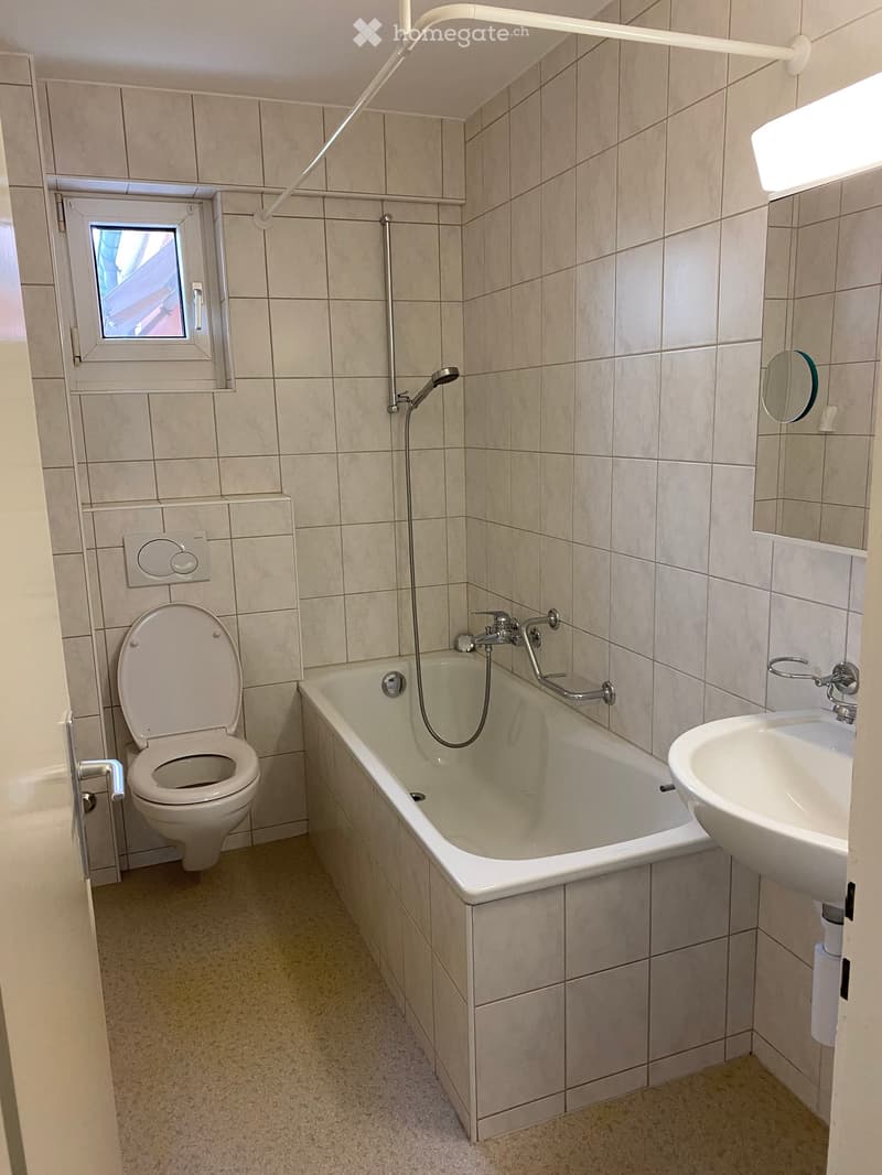 Bathroom with beige tiled walls and floor, featuring a toilet, bathtub with a handheld shower, a sink, and a small window above the bathtub.