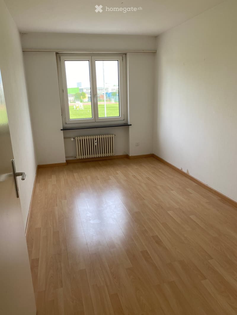 Empty room with light wooden flooring, white walls, a radiator, and a large window with a view of a green outdoor area.