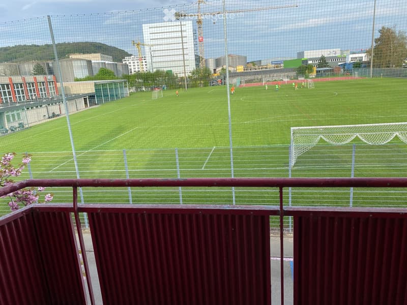 View of a fenced soccer field with players practicing, surrounded by buildings and construction cranes in the background.