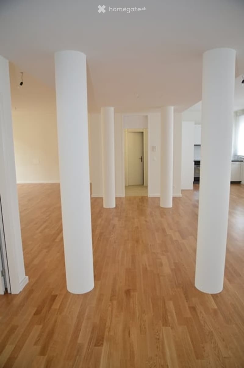 Empty room with light wooden flooring and white walls, featuring several white columns and open space.