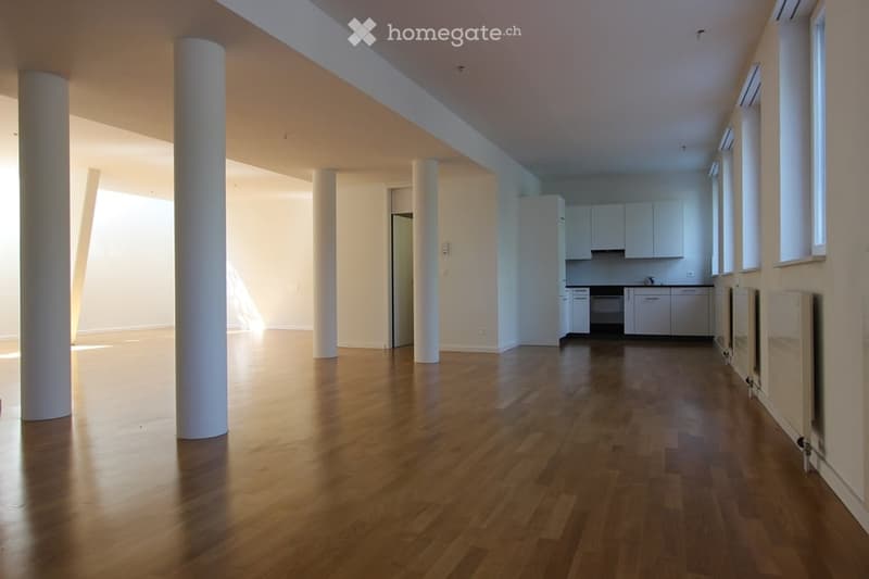 Open-plan unfurnished space with wooden flooring, white walls, and a small kitchen area featuring white cabinets and a sink.