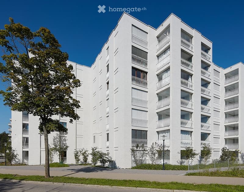 Modern white apartment building with multiple floors, balconies, and large windows. The building is surrounded by greenery and a paved area.