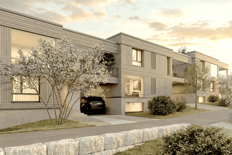 Modern two-story house with wooden siding, large windows, and a raised ground floor. Features a garage, balcony, and landscaped garden with trees and shrubs.