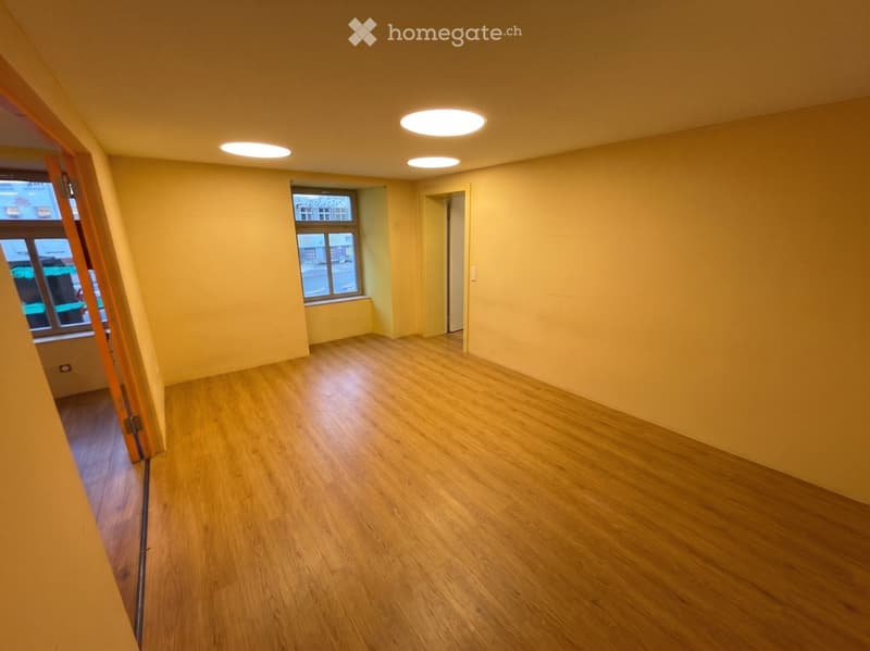Empty room with wooden flooring, light yellow walls, and recessed ceiling lights. There are windows and a doorway leading to another space.