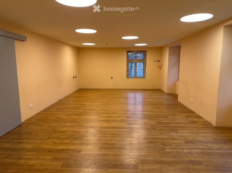 Empty room with wooden flooring, light-colored walls, and recessed ceiling lights. A single window provides natural light.