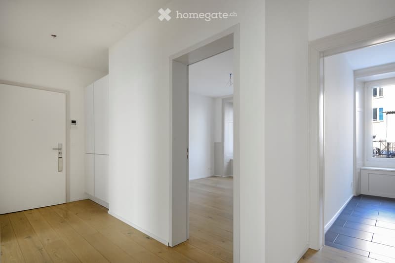 A bright, minimalist hallway with white walls, wooden flooring, and a partially open door leading to another room. The hallway features a sleek white door with a modern handle and a small intercom system mounted on the wall.