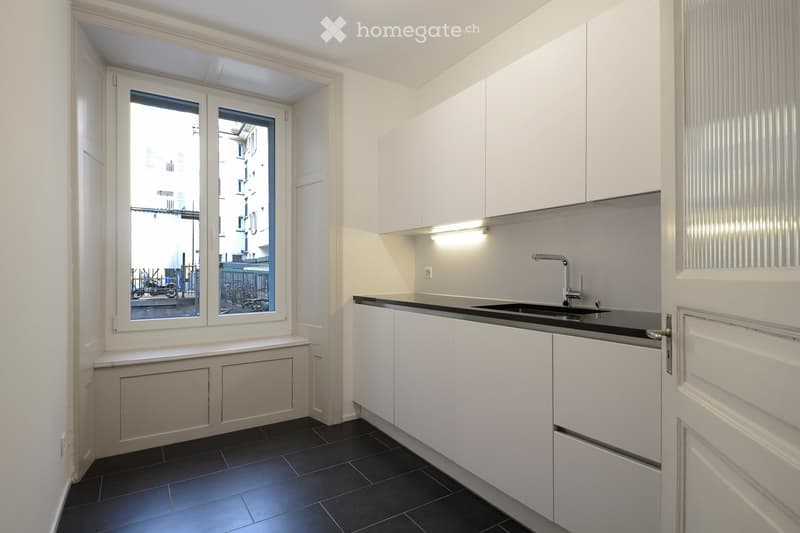 Modern kitchen with white cabinets, black countertop, and a sink with a modern faucet. The room features dark tiled flooring and large windows offering a view of the street.