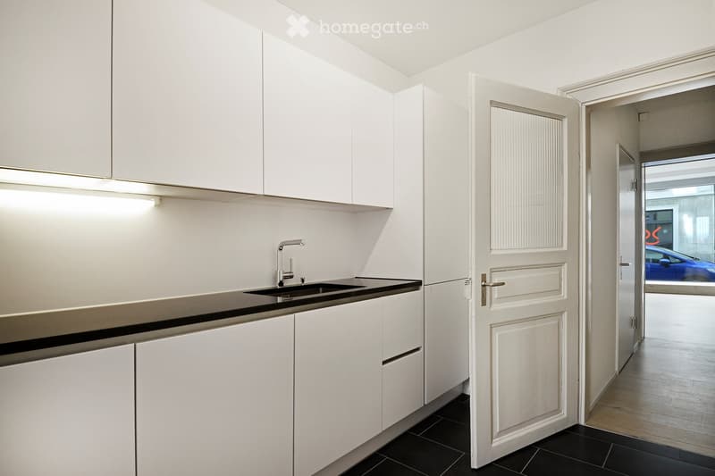 Modern kitchen with white cabinets, black countertop, and a sink with a chrome faucet. The floor is black tiled, and the space is well-lit with built-in lighting above the cabinets.