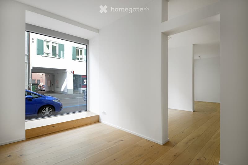 Empty room with light wooden flooring and white walls, featuring a large window with a view of the street and a parked car.