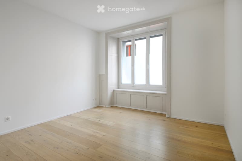 Empty room with light wooden flooring, white walls, and large windows with white frames. No furniture or equipment is present.