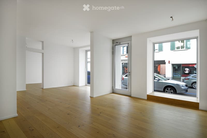 Empty room with white walls and wooden flooring, featuring large windows and a glass door leading outside.