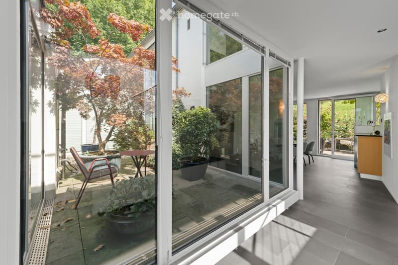 Garden / Outdoor Area. A modern, well-maintained outdoor area with large glass sliding doors leading to an indoor space. The patio features potted plants, a small table, and a red chair, with a lush garden visible through the glass.