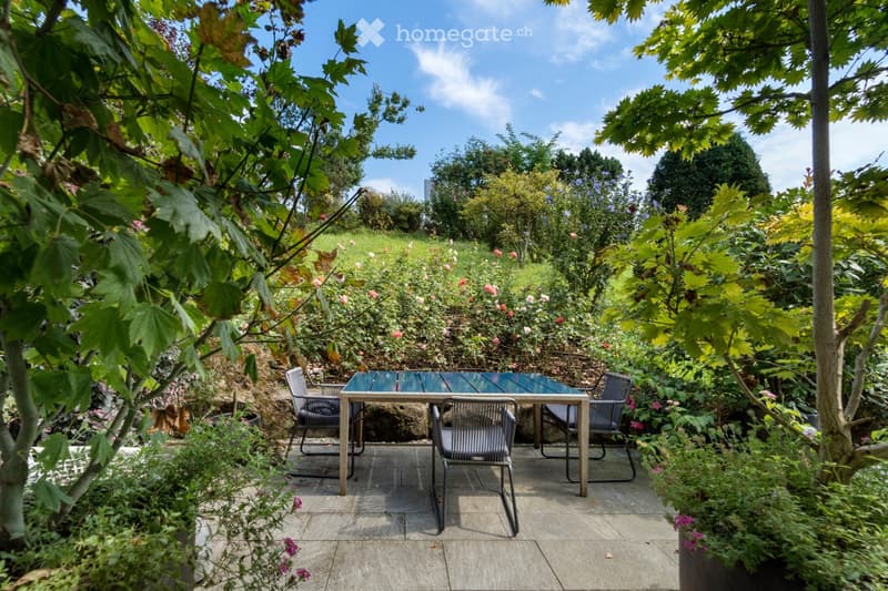 Garden / Outdoor Area. A lush garden with a patio area featuring a table and four chairs surrounded by greenery and flowers.
