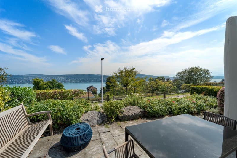 Garden / Outdoor Area. A sunny outdoor patio with a wooden bench, a black table, chairs, and a blue stool, overlooking a scenic lake and distant hills.
