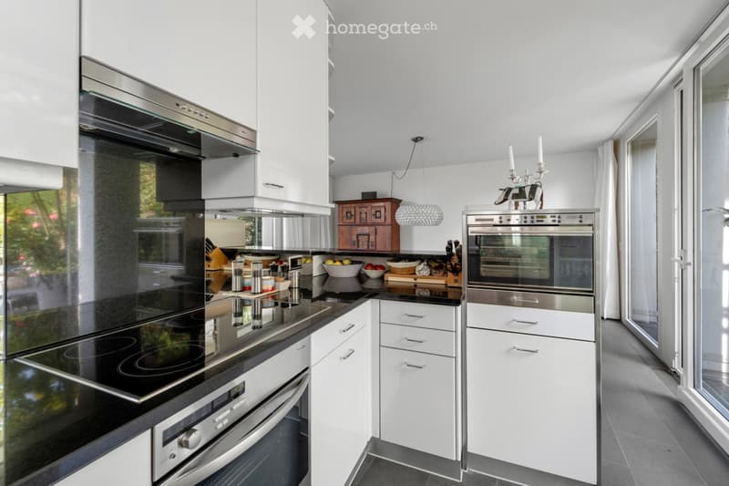 Kitchen. Modern kitchen with white cabinets, stainless steel appliances, induction stove, oven, and a small wooden cabinet. The countertop features a bowl of fruit and a cutting board with kitchen utensils.