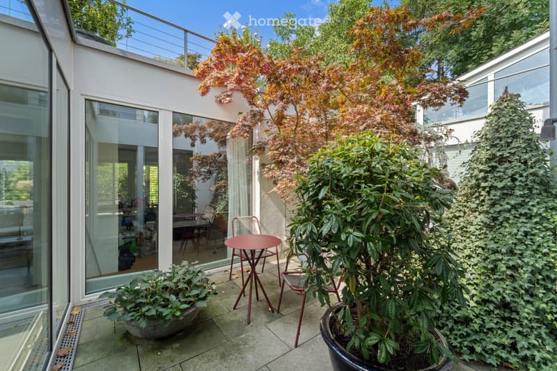 Garden / Outdoor Area. A modern outdoor patio area with a round red table and chairs, surrounded by potted plants and trees, with a glass-walled building in the background.