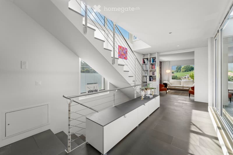 Corridor / Hallway. Modern hallway with a sleek staircase featuring a glass railing, white walls, and a built-in bench with a black countertop. The hallway leads to a living area with a sofa, coffee table, and large windows offering a view of greenery.