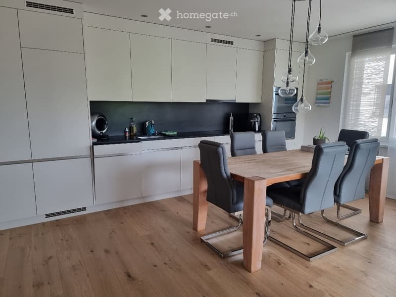 Modern kitchen and dining area with white cabinets, wooden dining table, grey chairs, pendant lights, and stainless steel appliances.