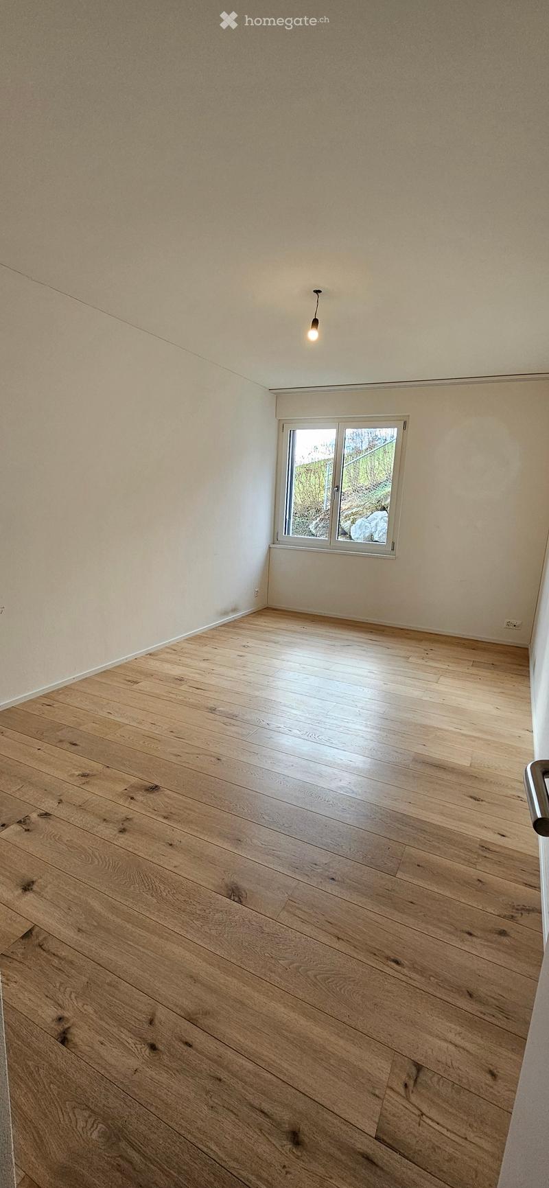 Empty room with wooden flooring, white walls, and a single window with a view of greenery outside.