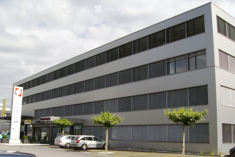 2 story office building, grey walls, multiple glass windows, entrance with awning, parking lot, cars parked, plants in front