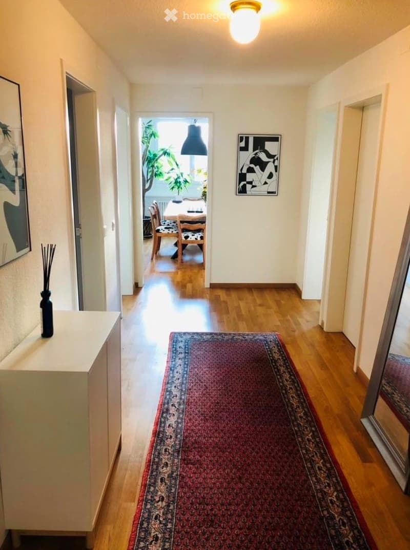 A well-lit hallway with wooden flooring, a patterned rug, and artwork on the walls. A small cabinet with a vase is visible on the left, and a dining area can be seen at the end of the hallway.