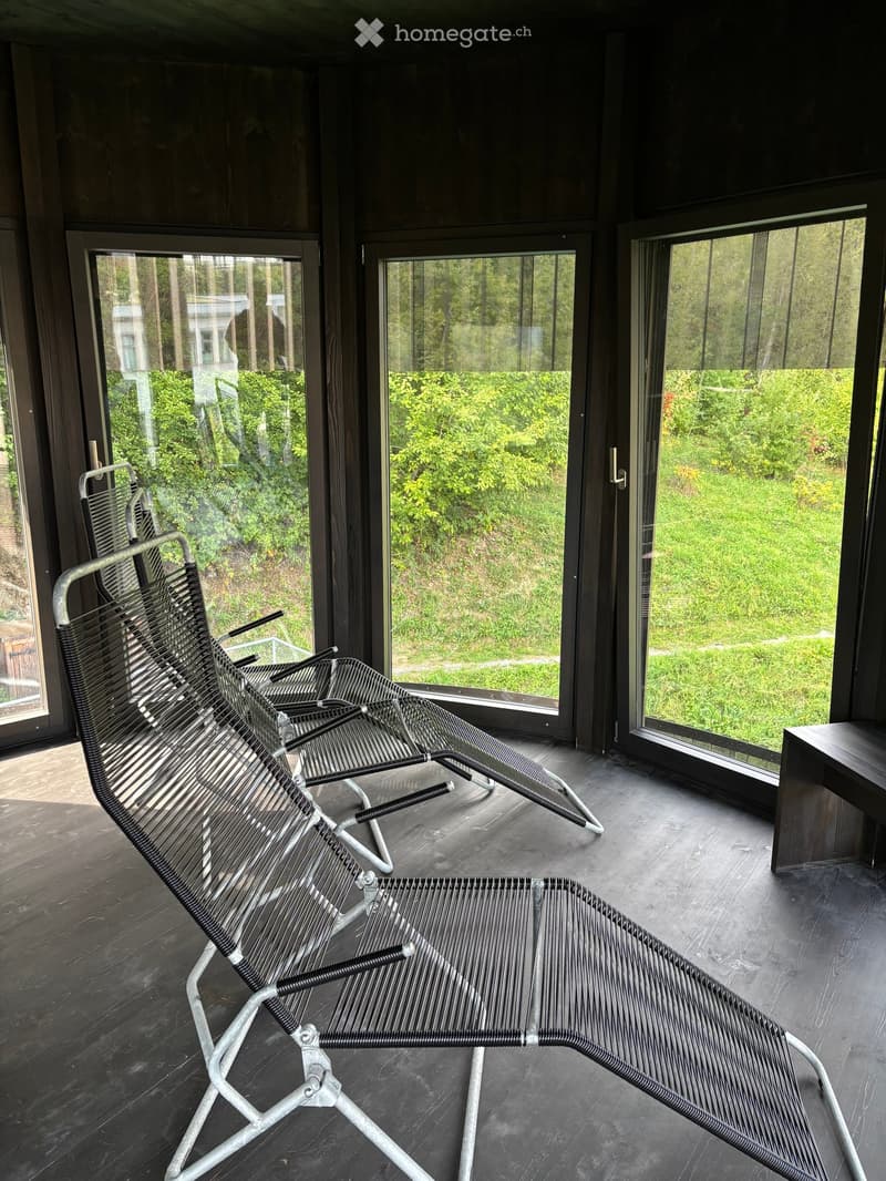 Modern balcony with large glass windows offering a view of greenery, featuring two metal lounge chairs and a small wooden table.