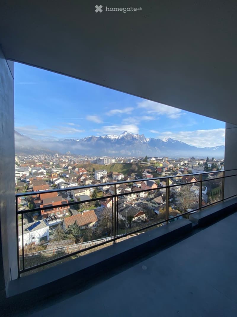 Balcony with a modern glass railing, overlooking a town with houses and distant snow-capped mountains.