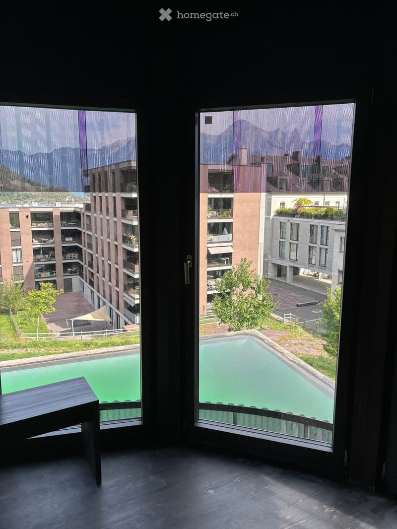 Modern room with large windows offering a view of a swimming pool, greenery, and surrounding buildings.