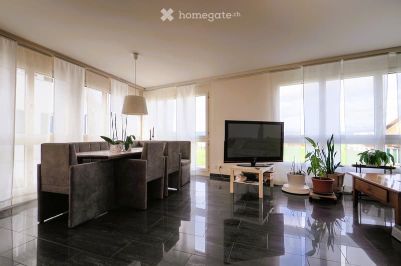 Modern living room with large windows, dark tiled floor, grey sofa, wooden coffee table, TV, and potted plants.