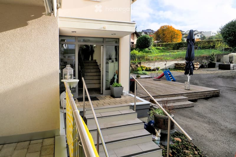 Outdoor area with wooden deck, steps leading up to a house entrance, potted plants, a small table with a lantern, and a closed umbrella on the deck.