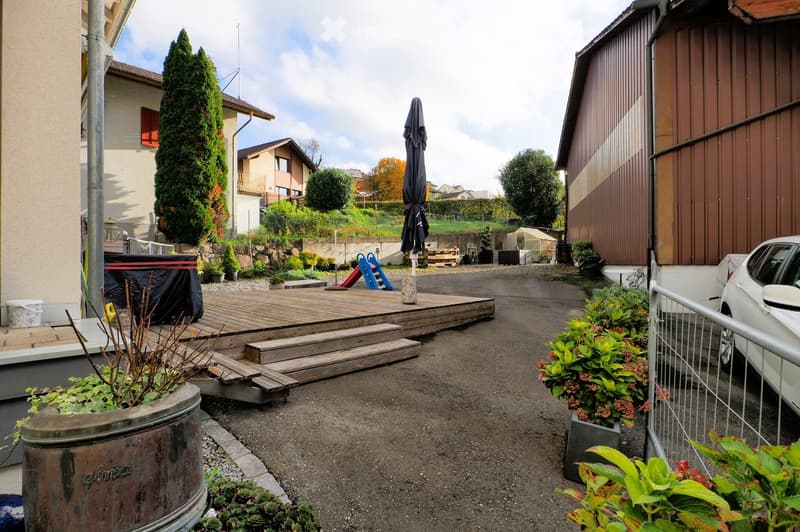 Outdoor area with a wooden deck, steps leading up, potted plants, a covered hot tub, a slide, and a closed umbrella. A white car is parked nearby, and there are buildings and trees in the background.