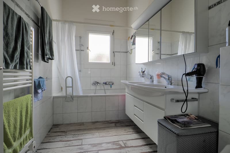 Modern bathroom with a bathtub, double sink, mirror, and wooden flooring. Includes towels, a hair dryer, and a small stool with a magazine.