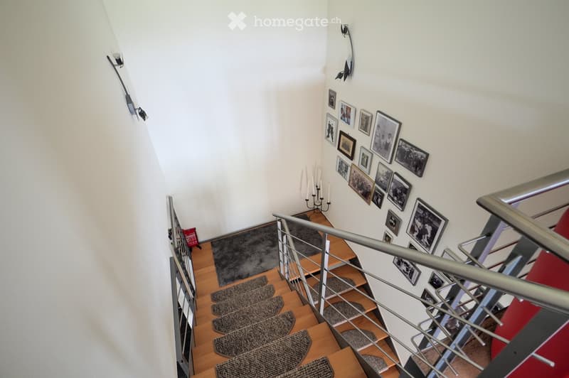 Spiral staircase with wooden steps, metal railing, and framed pictures on the wall. The staircase features a small landing area with a candle holder and a red bag on the floor.