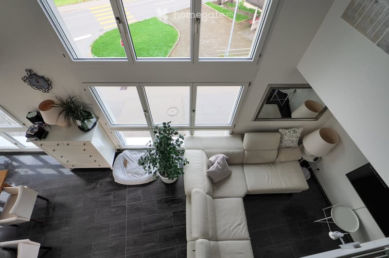 Modern living room with large windows, white sectional sofa, grey throw pillows, a white armchair, a small round table, a potted plant, and a lamp. The room features dark grey tiled flooring and white walls.