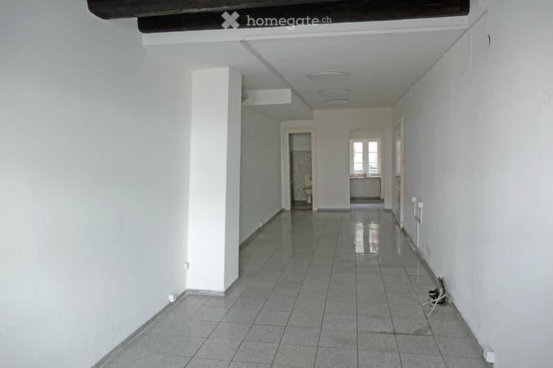 Empty hallway with white walls, tiled floor, and several doors leading to rooms, including a visible bathroom with a toilet and sink.