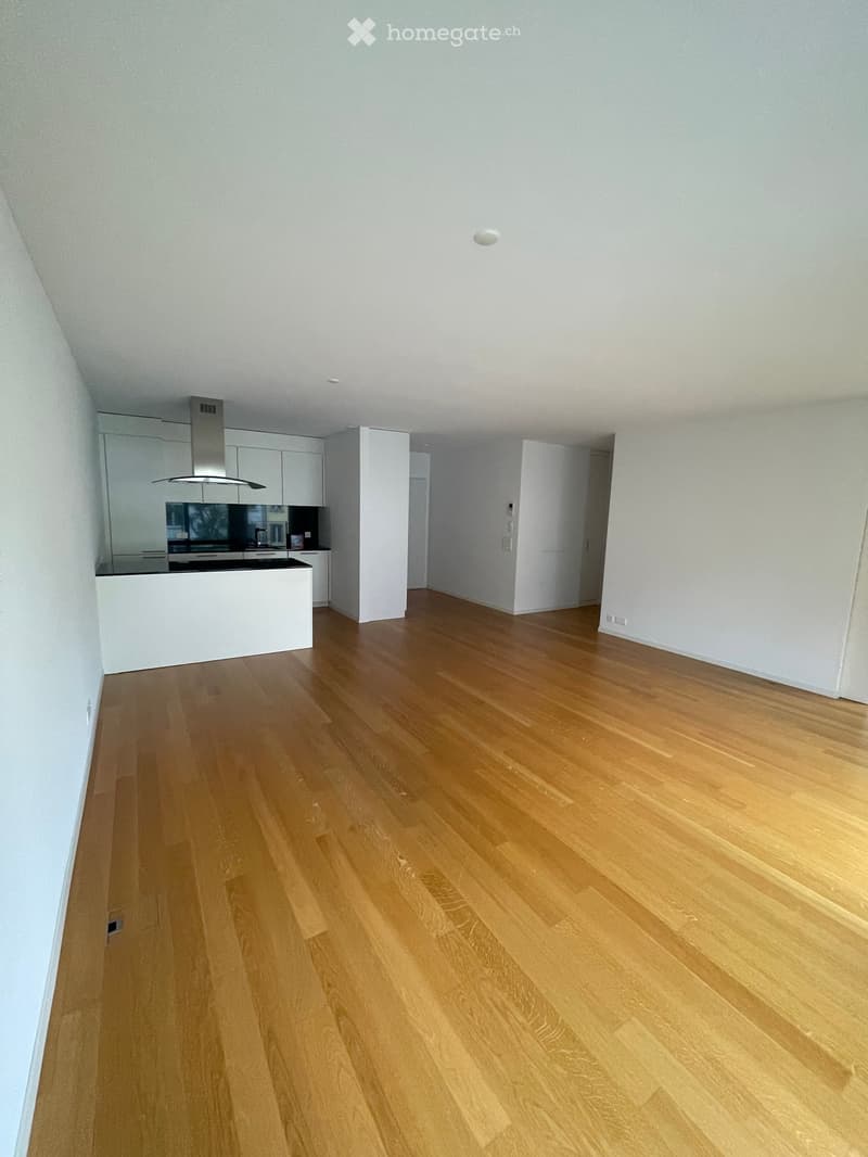 Empty room with light wooden flooring and white walls, featuring a modern kitchen area with cabinets and a countertop in the background.