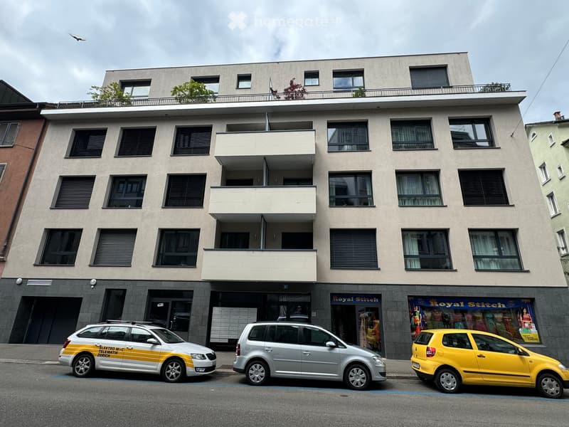 Modern multi-story building with balconies, large windows, and a rooftop terrace. Three cars are parked in front.