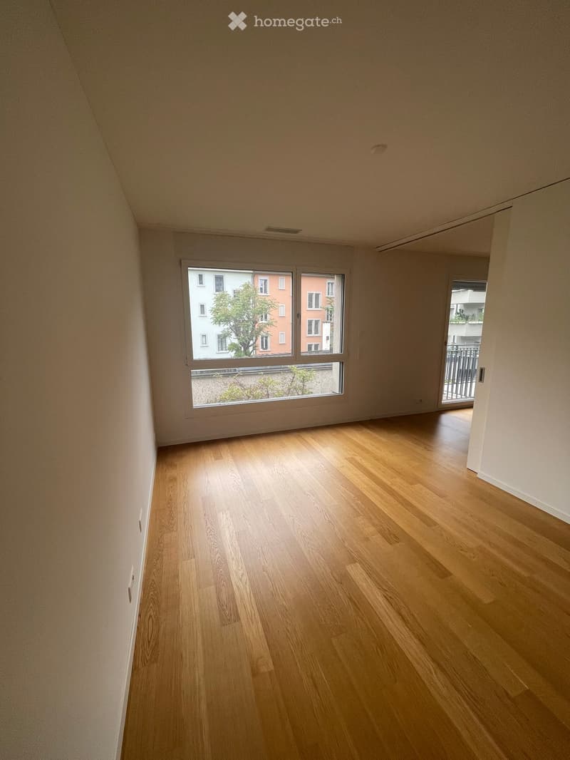 Empty room with light wooden flooring and large windows offering a view of the city and a balcony area.