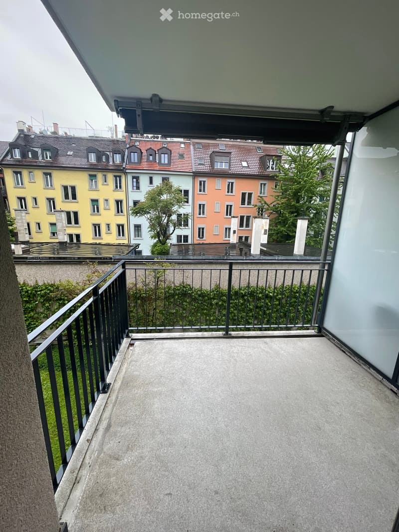 Balcony with concrete floor, black metal railing, and view of surrounding buildings and greenery.