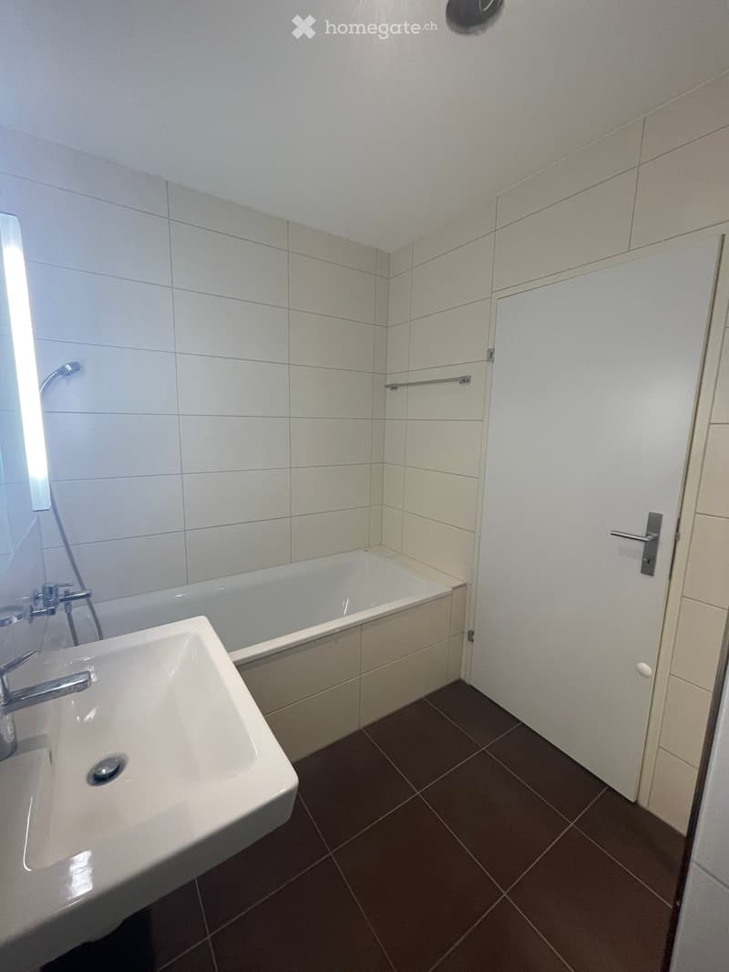 Modern bathroom with white tiled walls, a white sink, a bathtub, and a door with a silver handle.