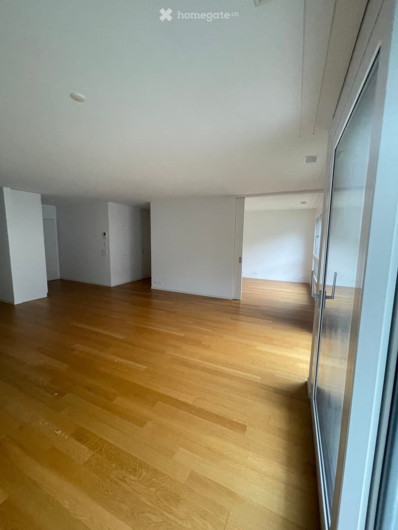 Empty hallway with light wooden flooring and white walls, featuring several doors and a glass partition on the right side.