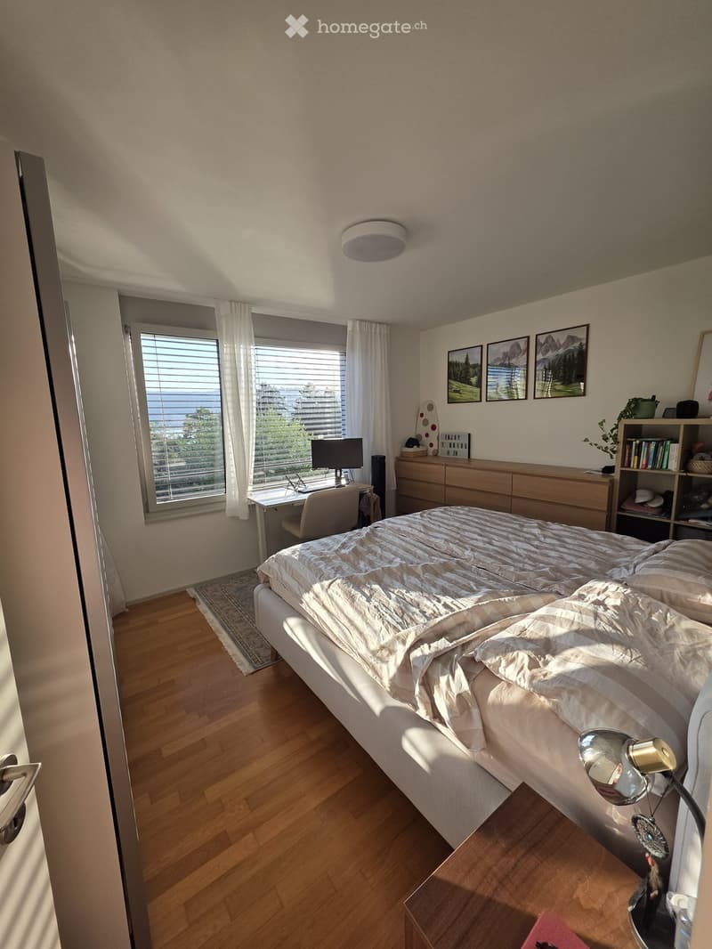 Modern bedroom with a large bed, nightstand, desk, and large window with blinds. The room features wooden flooring and several decorative items including picture frames and a shelf.
