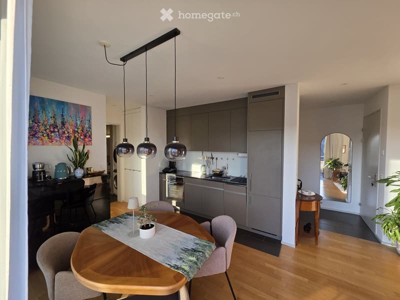 Modern kitchen and dining area with grey cabinets, wooden flooring, and pendant lights. Includes a round wooden dining table with chairs, a small bar area, and a kitchen island with appliances.