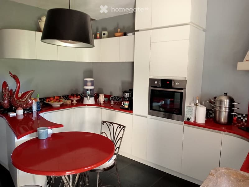 Modern kitchen with white cabinets, red countertops, a round red dining table with a single chair, an oven, a water dispenser, and various kitchen items on the counters.