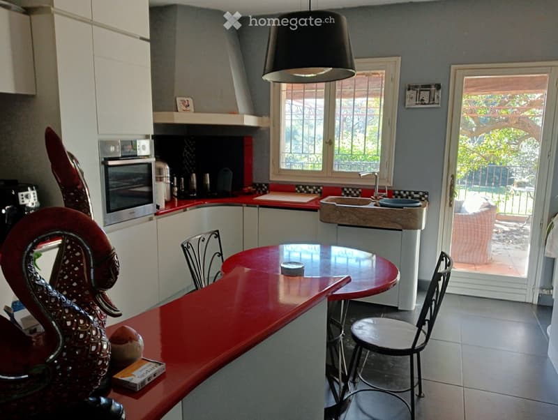 Modern kitchen with red countertops, white cabinets, a sink, an oven, and a dining area with a round table and chairs. A large window and a sliding glass door lead to a balcony.