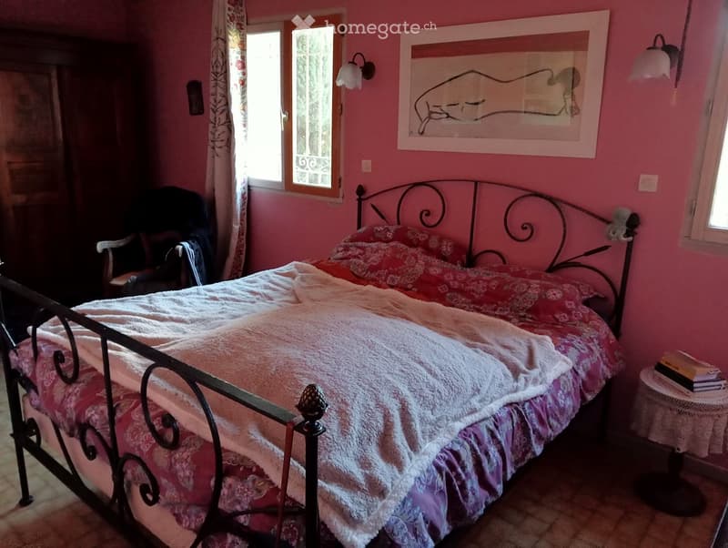 Pink bedroom with a metal-framed bed, floral bedding, a wooden chair, a small table with books, and a framed picture on the wall.