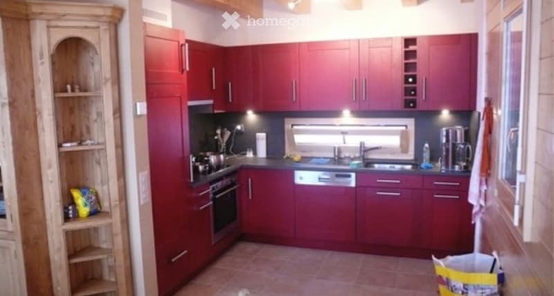 A modern kitchen with red cabinets, a sink, an oven, a dishwasher, and a coffee maker. The space is well-lit with built-in lighting and features a window above the sink.