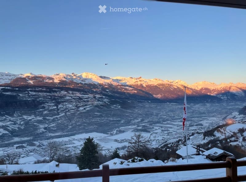 Panoramic view of snow-covered mountains and a town from a balcony, with a wooden railing and a flag visible.